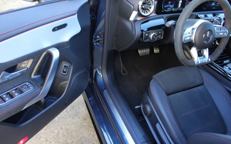 Vista del puesto de conducción desde la puerta abierta, mostrando el volante AMG, pedales deportivos y detalles en aluminio cepillado.